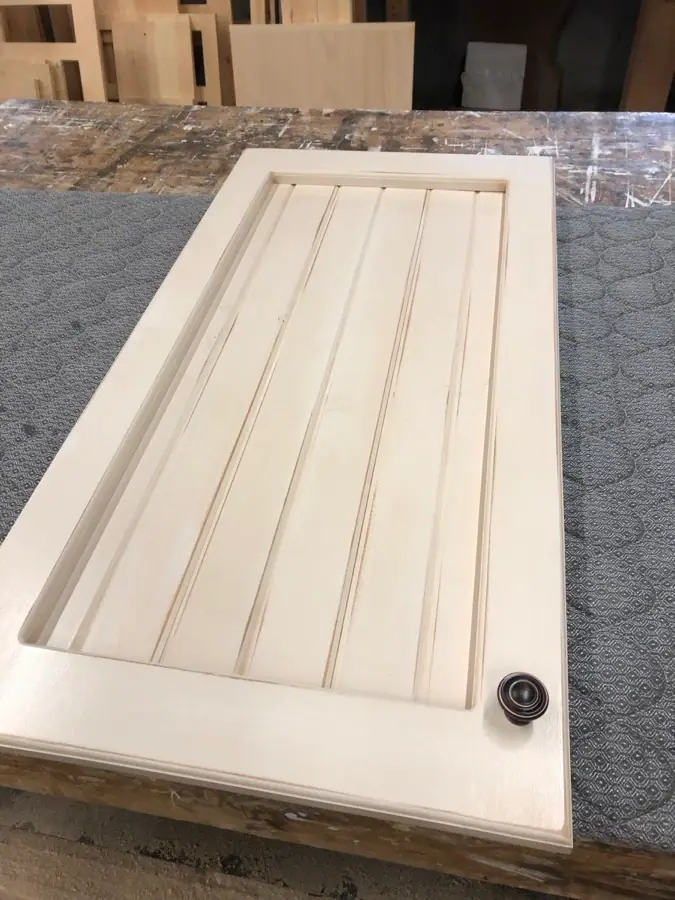 Porte de modèle lambris en pin, peinte blanc crème avec bouton noir, posée sur l’établi de l’atelier.